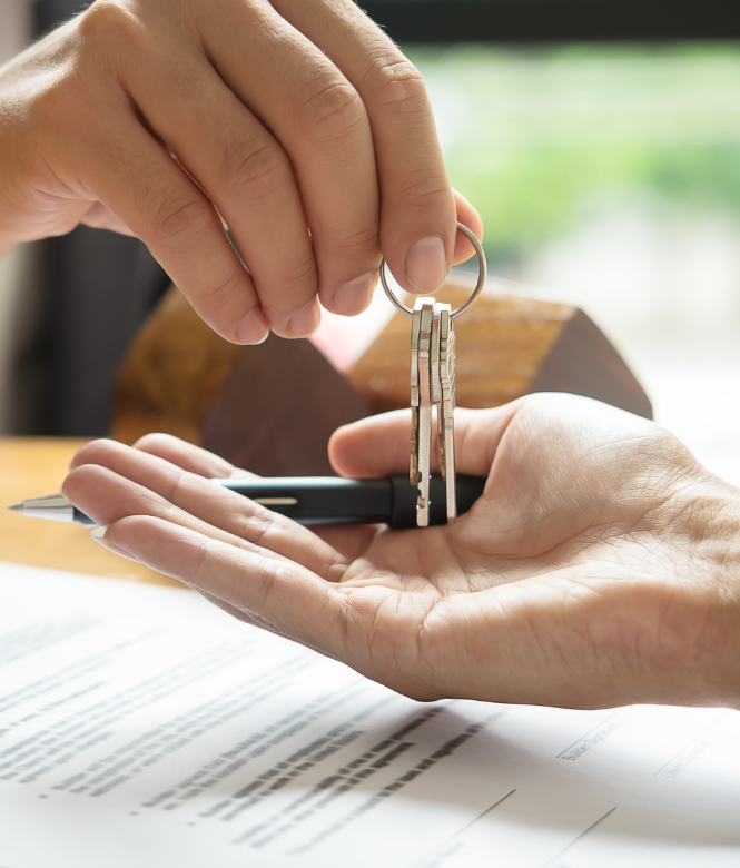 A person handing over house keys above a contract with a small house model in the background.