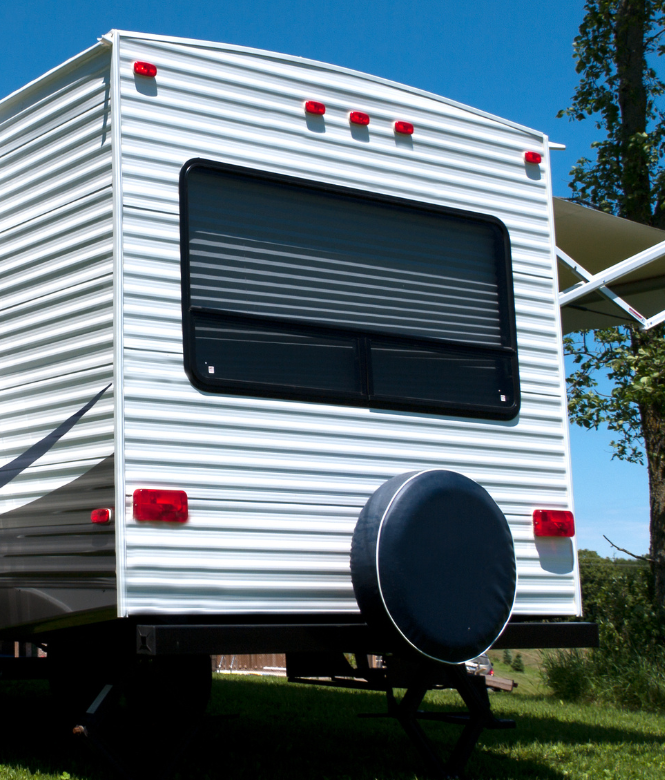Back of a white travel trailer with spare tire and large window under a sunny blue sky.
