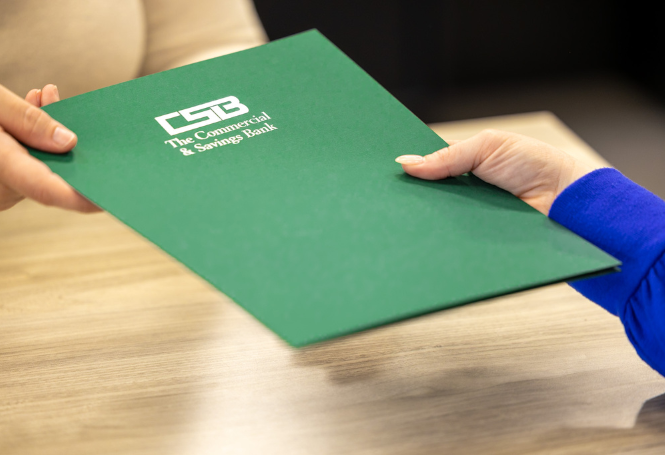 Hands exchanging green CSB folder at bank desk.